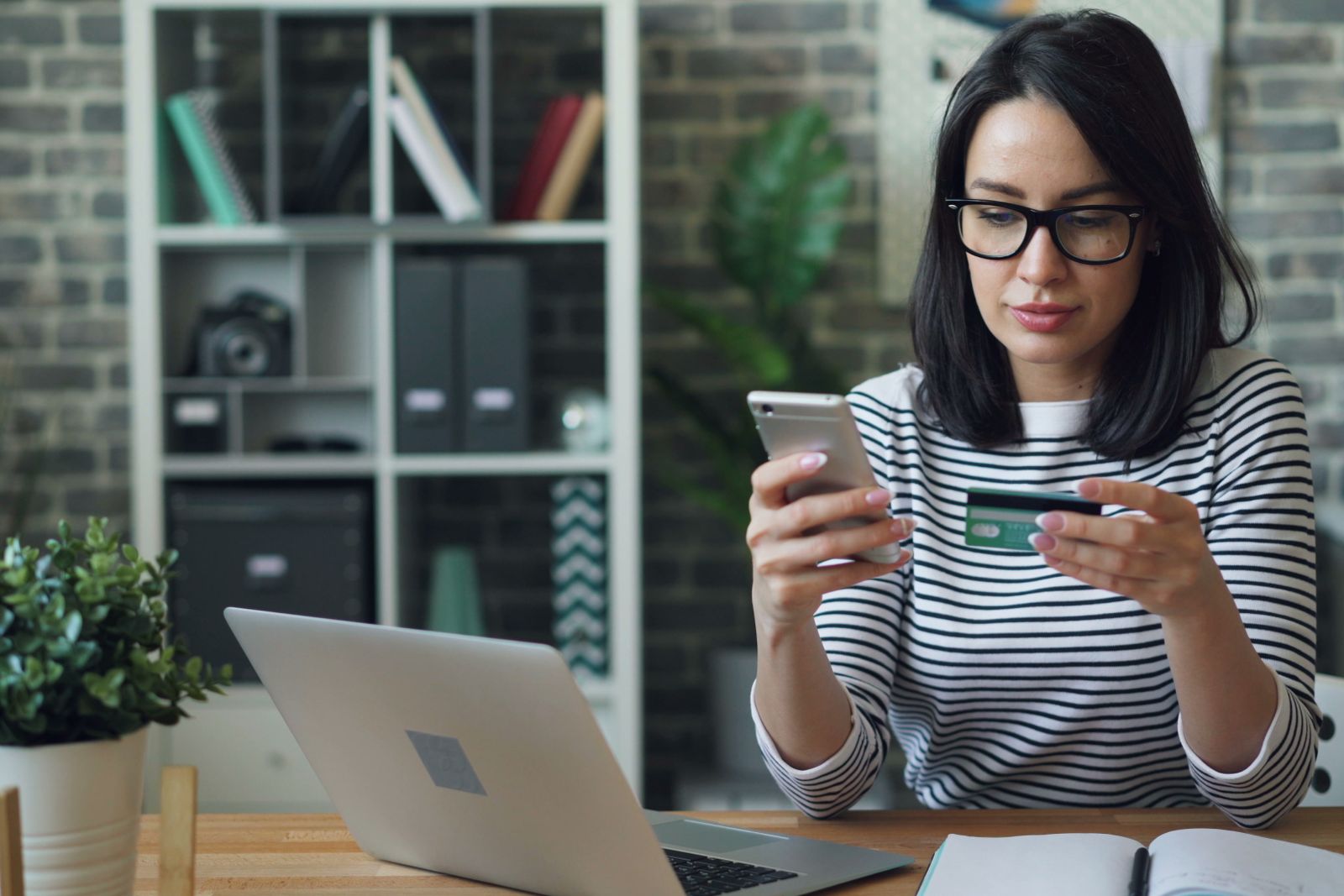 Entrepreneur smiling at a payment card while checking her phone beside a laptop